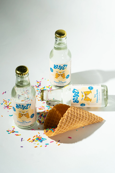 Three clear glass bottles of Burp! Ice Cream Soda with gold caps and colorful sprinkles, arranged around an open waffle cone on a white background.